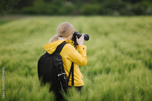 Back view of professional photographer in yellow jacket taking photos of green wheat field during spring time. Mature lady with black backpack enjoying favorite hobby outdoors.