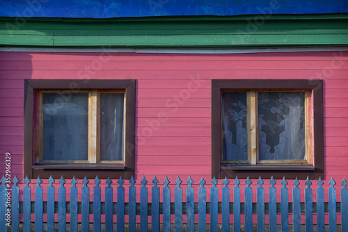 Two small windows in a wooden, pink-painted wall of a village house behind a blue fence