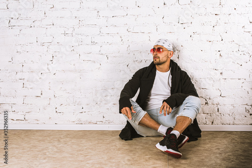 Pensive stylish young man in bandana and pink glasses sitting on the floor and looking at copyspace in a bright room 