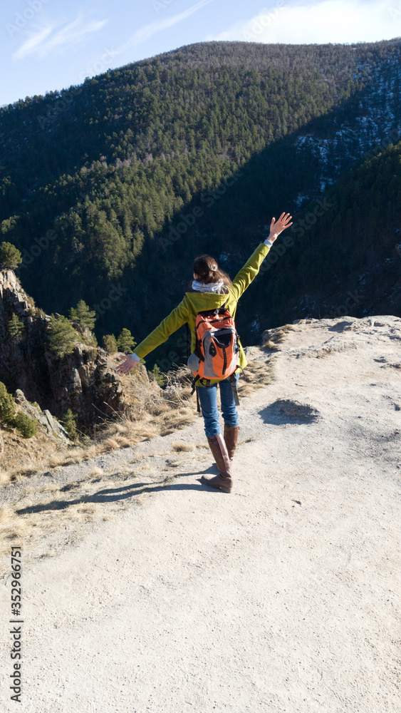 A female traveler in a coat and a backpack stands on the edge of a cliff. The vertical frame. Brunette hands raised to the side. Success