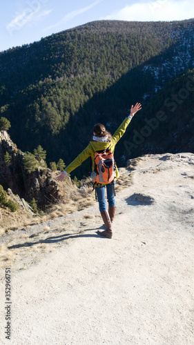 Obraz na płótnie A female traveler in a coat and a backpack stands on the edge of a cliff. The vertical frame. Brunette hands raised to the side. Success w salonie