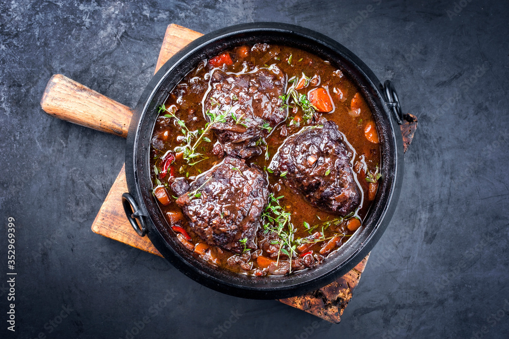Traditional German braised beef cheeks in brown red wine sauce with carrots and onion offered as top view in a modern design stewpot on an old rustic board - obrazy, fototapety, plakaty 