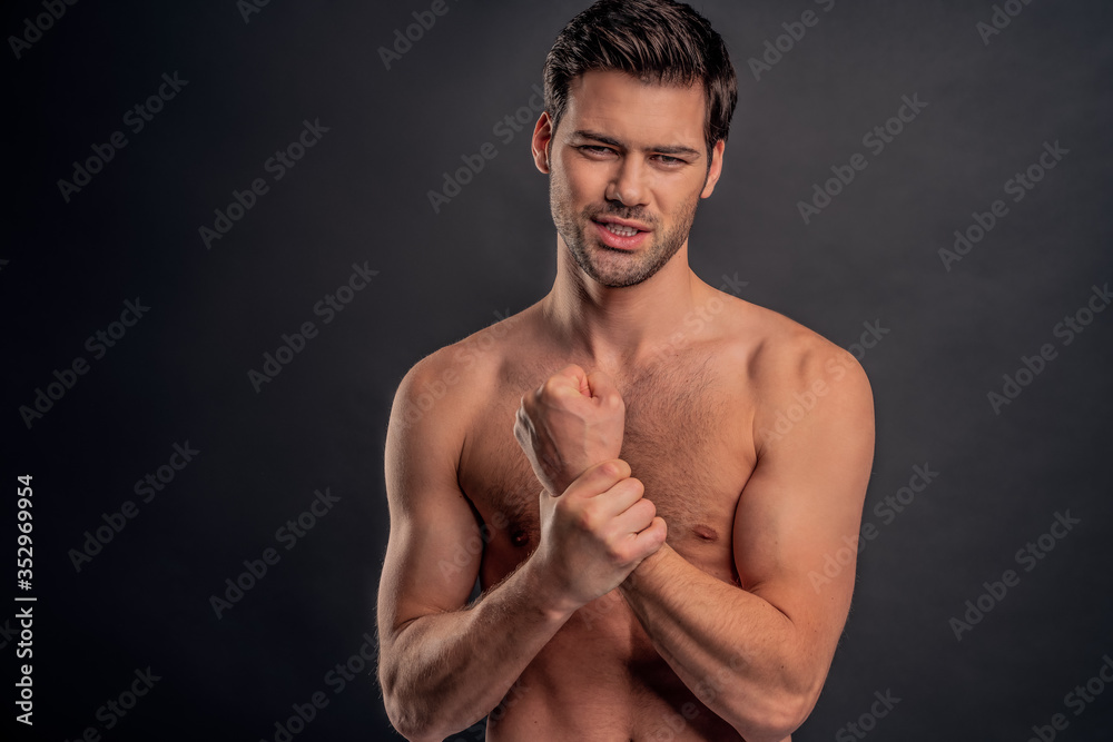 © Roman - Handsome young bearded man isolated. Cropped image of topless muscular man is standing on gray background. Man holding his wrist. Experiencing wrist pain.