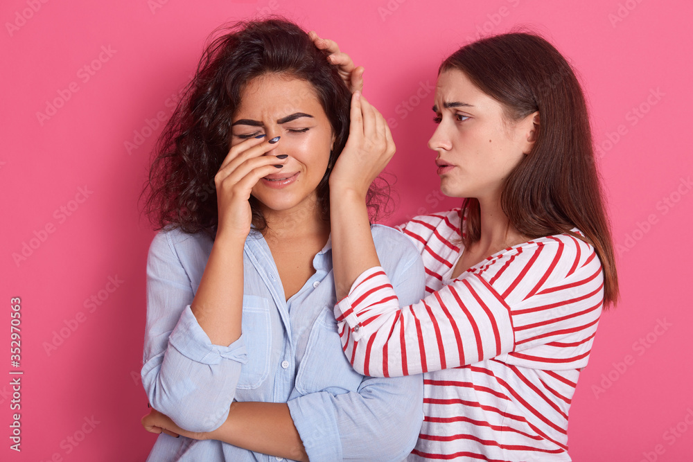 Two girlfriends standing against pink wall, desirable lady with dark hair comforts her crying friend, lachrymose female wears blue casual shirt being upset, suffering from unrequited love. Two girlfriends standing against pink wall, desirable lady with dark hair comforts her crying friend, lachrymose female wears blue casual shirt being upset, suffering from unrequited love.