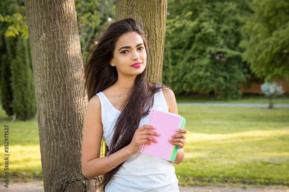 Young beautiful Indian girl stands in the park near a tree, holding a notebook in his hands. Portrait of a female student girl outdoors Young beautiful Indian girl stands in the park near a tree, holding a notebook in his hands. Portrait of a female student girl outdoors