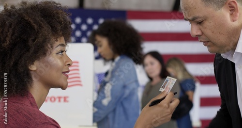 CU Mixed-race staff person inspects photo ID, driving license, presented by Hispanic man at US polling station. Other voters soft focus in background with US flag