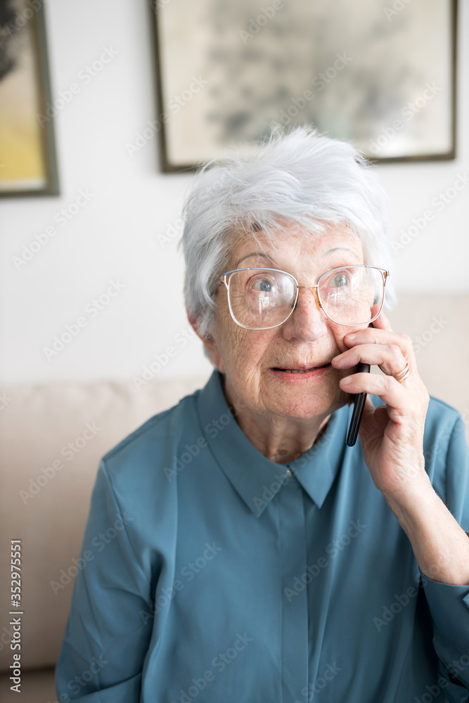 Smiling senior gray-haired Caucasian woman sitting on a sofa in brightly lit living room and talking on the phone Smiling senior gray-haired Caucasian woman sitting on a sofa in brightly lit living room and talking on the phone