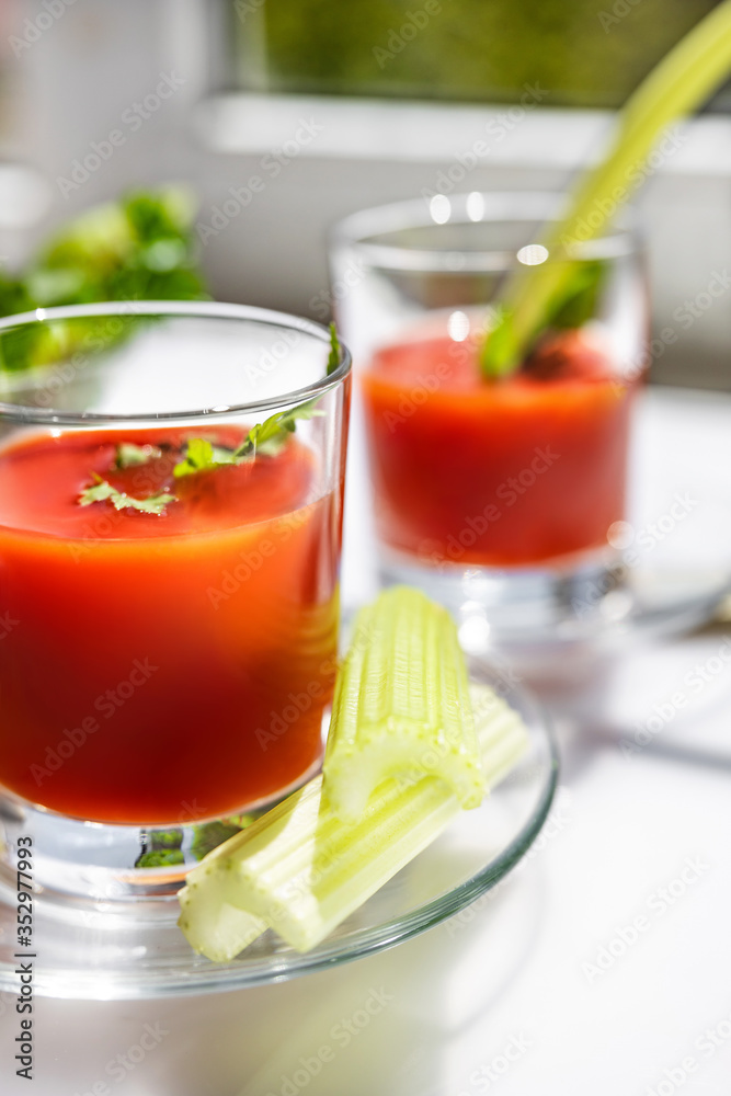 Two glasses of tomato juice with parsley and celery decorations, stand near the window, morning sunlight shines, shallow depth of field, selective focus. Natural drinks concept. Two glasses of tomato juice with parsley and celery decorations, stand near the window, morning sunlight shines, shallow depth of field, selective focus. Natural drinks concept.