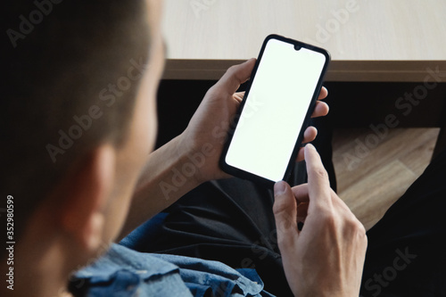 A businessman holds a mobile phone in his left hand and looks at the white screen of the mockup. Concept of online communication, work and technology
