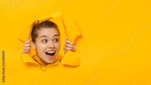 Horizontal banner of young female tearing paper and peeking out hole, curious about commercial offer on copy space on right, isolated on yellow background