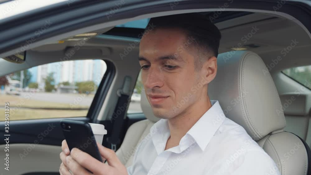 Portrait young man businessman sitting in a car, using smartphone and drinking coffee. Wearing a white shirt. Portrait young man businessman sitting in a car, using smartphone and drinking coffee. Wearing a white shirt.