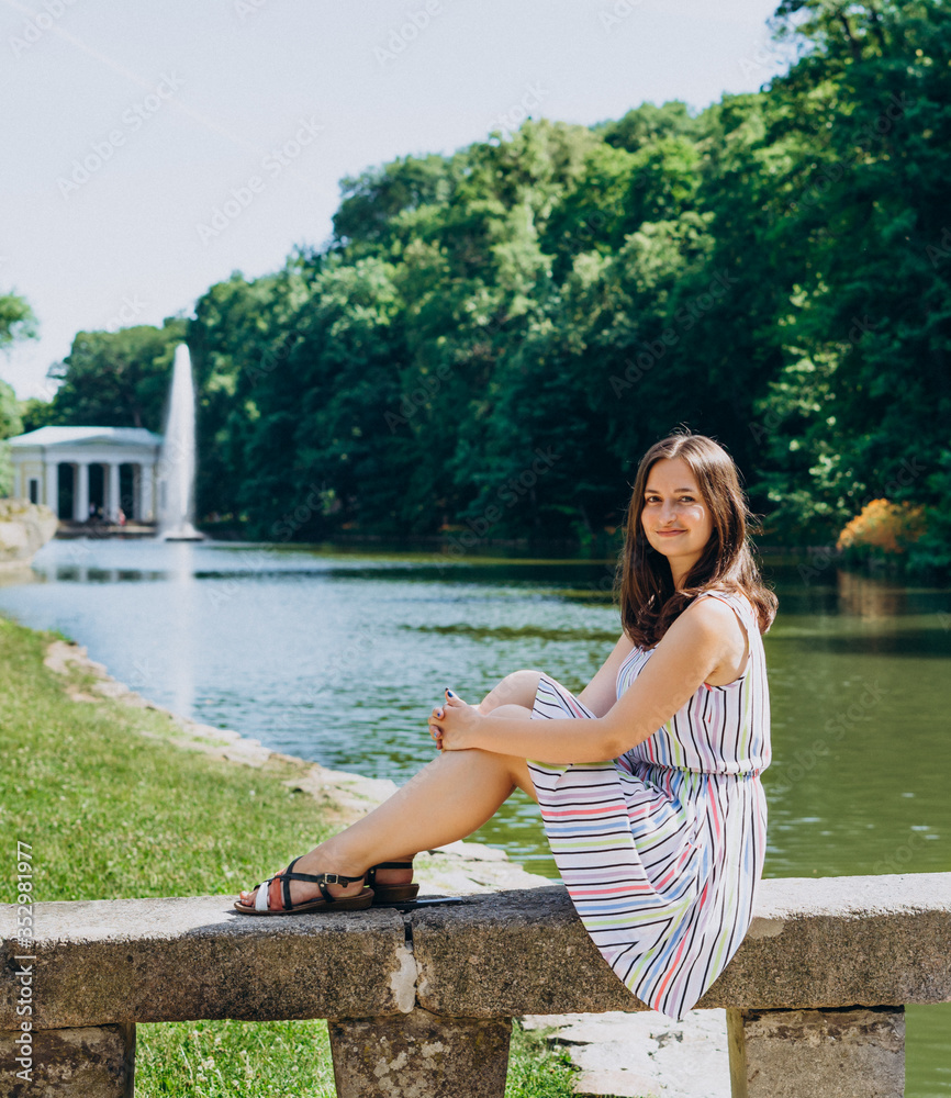 Sofia Park, Uman. Young woman sitting on a bench on the background of the lake with a fountain. The girl in a dress sits on a stone bench. Girl on a tour of the national park on a summer day. Sofia Park, Uman. Young woman sitting on a bench on the background of the lake with a fountain. The girl in a dress sits on a stone bench. Girl on a tour of the national park on a summer day.