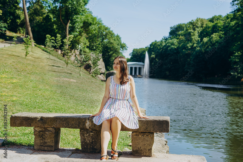 Naklejka premium Sofia Park, Uman. Young woman sitting on a bench on the background of the lake with a fountain. The girl in a dress sits on a stone bench. Girl on a tour of the national park on a summer day.