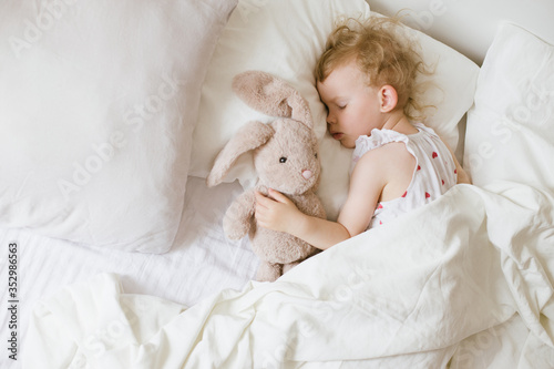 Cute child little girl sleeping in bed with white linen hugging her soft toy teddy bunny.