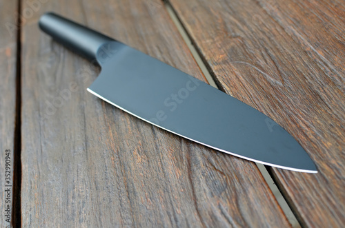 Close-up of a kitchen knife on wooden background with a black handle and black blade, matte finishing, flat lying, perspective view, focus on foreground