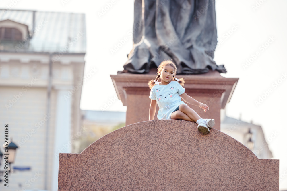 Obraz premium Portrait of a lovely little girl siiting and having fun on the monument in park.