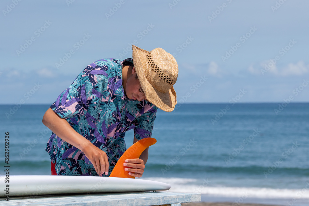 Fototapeta premium Putting a surfboard fin into a surfboard, the fin is orange in color and there is an Asian boy wearing a Hawaiian shirt and straw hat with red shorts. Chiba, Japan Surfing In Japan, Japan Surf