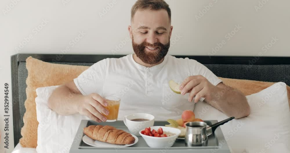 Young smiling man sitting with tray full of food on his legs and enjoing morning. Handsome guy eating apple and holding glass of juice while having breakfast in bed. Home background. Young smiling man sitting with tray full of food on his legs and enjoing morning. Handsome guy eating apple and holding glass of juice while having breakfast in bed. Home background.
