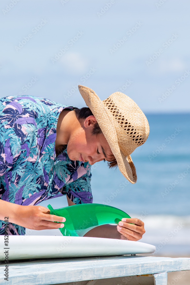 Fototapeta premium Surfer putting a green fin into a white surfboard on the beach with a blue ocean in the background he is Asian and wearing Hawaiian shirt and straw hat. He is also has a suntan