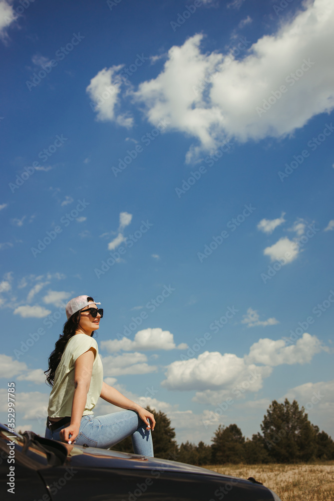 Obraz premium Outdoor adventure. Young woman sitting on the car hood while traveling. Tourism lifestyle, vacation, freedom and travel concept