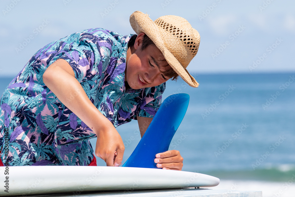 Fototapeta premium Japan Surf a boy putting a bluefin into a surfboard on the beach in Chiba close to the Olympic Surfing venue. He is wearing a Hawaiian shirt and straw hat & has a suntan. he is Japanese & has a smile.
