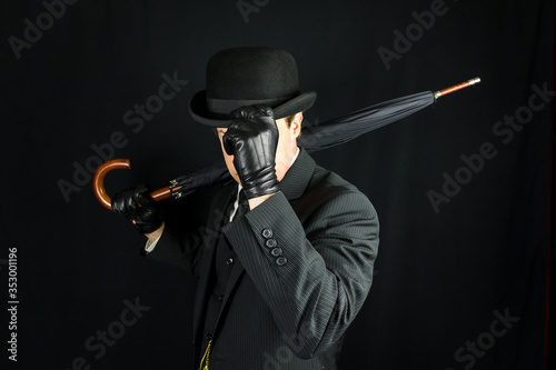 Portrait of British Businessman in Dark Suit and Bowler Hat Holding Umbrella. Hat Tipping Gesture. Concept of Classic and Eccentric English Gentleman. Film Noir Spy Hero photo