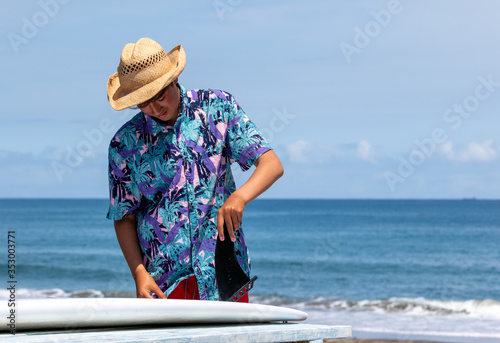 Black Surfboard fin being put onto a white surfboard by an Asian teenage surfer wearing a Hawaiian shirt and cool straw hat on a sunny beach with a blue ocean in Chiba, Japan.