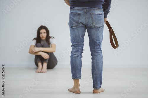 Scared woman hides near the wall while her husband swings with a belt.