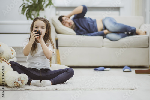 Drunk daddy laying on white sofa and watching how his little daughter tastes alcohol.