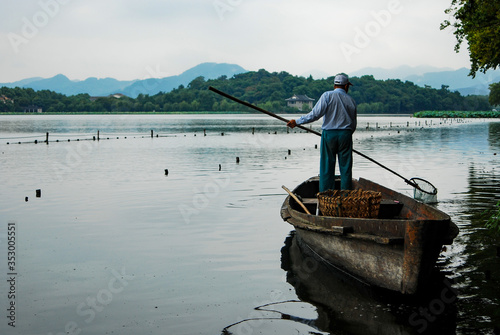 An elderly man standing on the boat cleaning the lake with a long stick and net at the end.