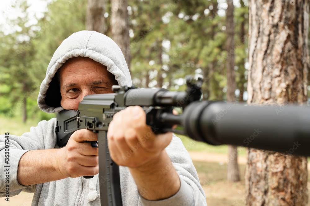 Obraz premium Front view of caucasian man wearing hood holding rifle semi automatic gun with silencer in the woods aiming ready to shoot or kill terrorist assassin murderer aim shooting in nature sunny day