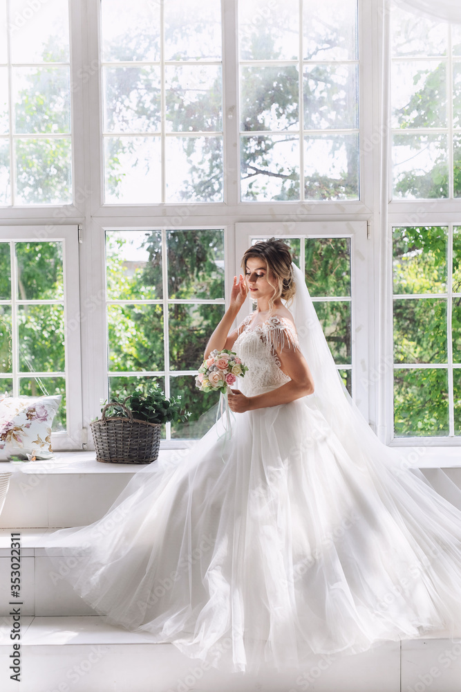 A gorgeous bride in a beautiful white dress with a bouquet of pink flowers sitting on the opposite side of a large window with a green lush garden outside.The bride is waiting for the groom. A gorgeous bride in a beautiful white dress with a bouquet of pink flowers sitting on the opposite side of a large window with a green lush garden outside.The bride is waiting for the groom.
