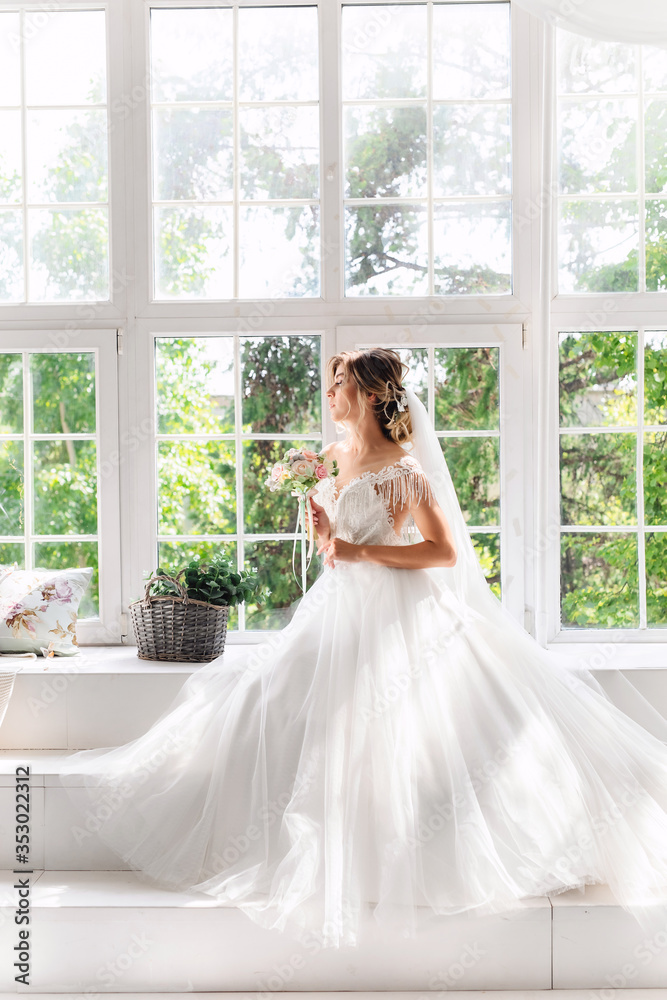 A gorgeous bride in a beautiful white dress with a bouquet of pink flowers sitting on the opposite side of a large window with a green lush garden outside.The bride is waiting for the groom. A gorgeous bride in a beautiful white dress with a bouquet of pink flowers sitting on the opposite side of a large window with a green lush garden outside.The bride is waiting for the groom.