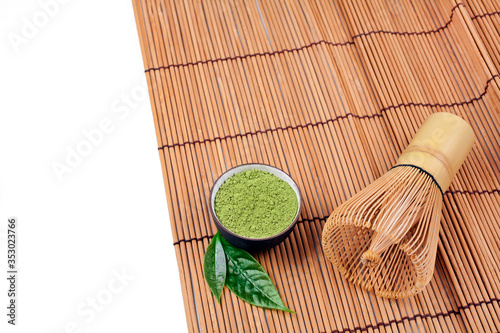Heap of powdered matcha tea in a clay cup and green leaves with bamboo matcha tea whisk also know as chasen on a bamboo background.