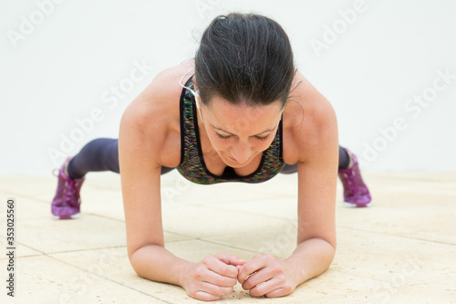 Focused woman with earphones standing in plank and looking at floor. Caucasian middle-aged lady in sport bra and leggings training alone. Sport and fitness during quarantine concept