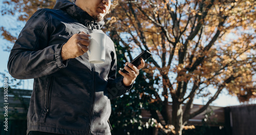 Asian man Standing at backyard using mobile phone and drinking coffee. Man relaxing at home reading text message on his cell phone in morning.