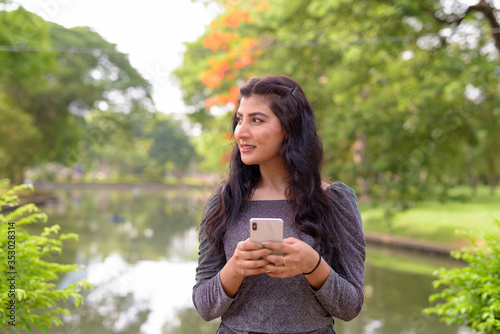 Happy young beautiful Indian woman thinking while using phone at the park outdoors