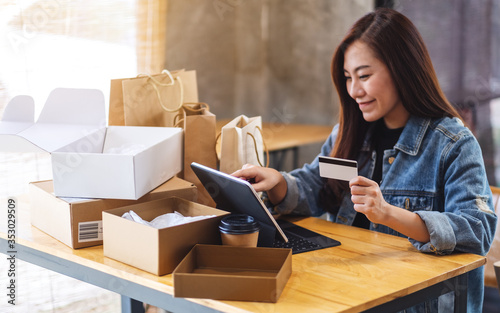 Closeup image of a young asian woman using tablet pc and credit card for online shopping with postal parcel box and shopping bags on the table