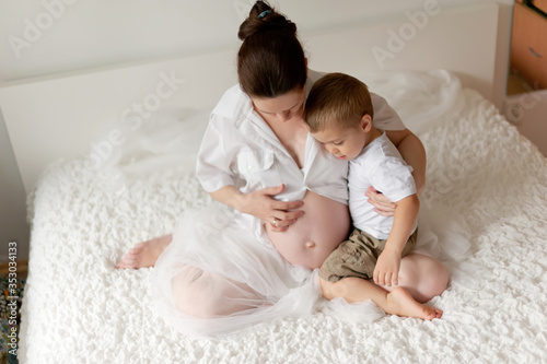 Active pregnant woman sitting on bed and touching her belly at home, with her little son hugs belly. A pregnant smiling girl is resting with her family on a white bed. The last months of pregnancy. photo