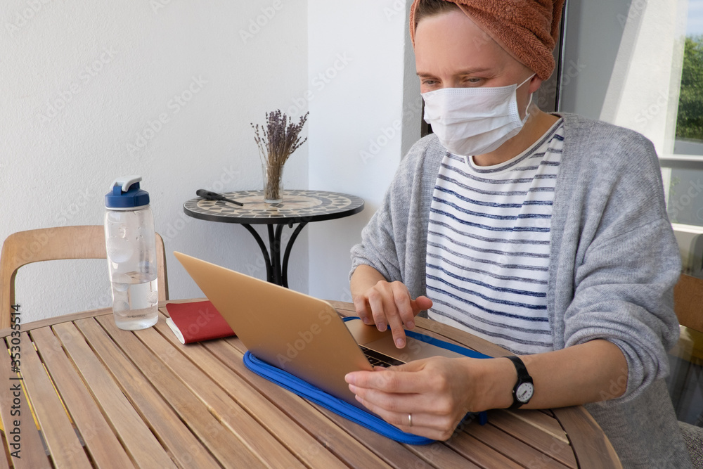 Woman in medicine mask working with laptop. Young lady sitting at round wooden table and working at home on self-isolation. Online job during quarantine concept Woman in medicine mask working with laptop. Young lady sitting at round wooden table and working at home on self-isolation. Online job during quarantine concept