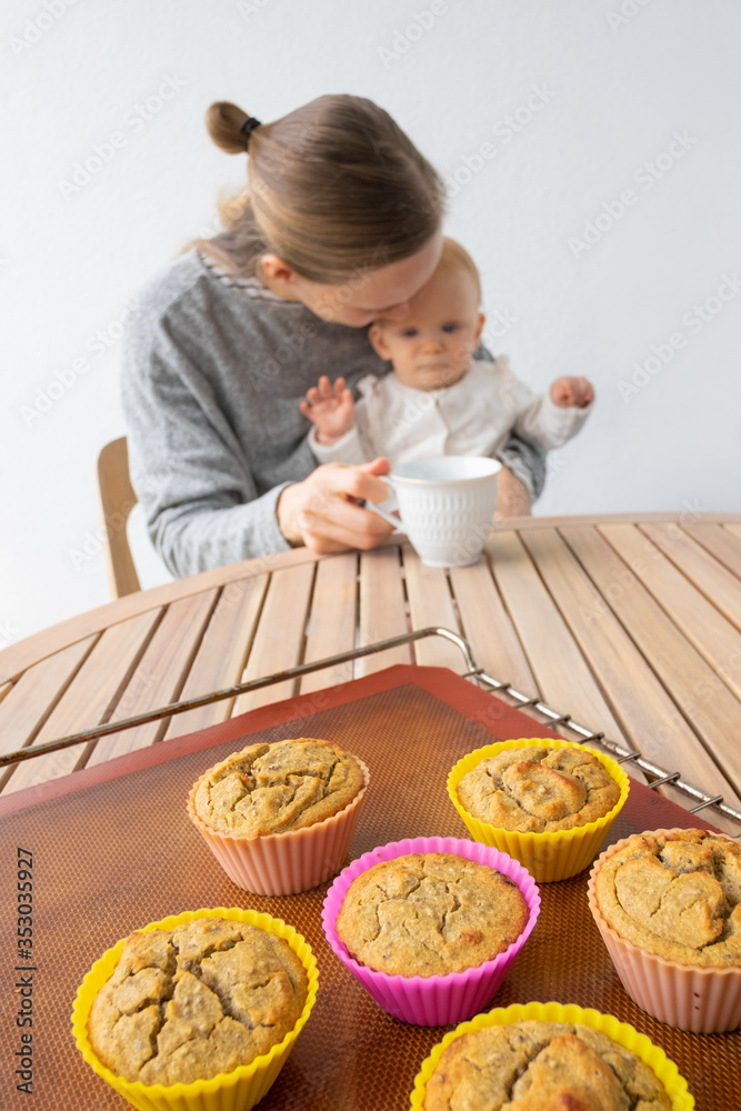 New mom holding baby in arms and drinking tea at table with freshly baked cupcakes. Child care and home baking concept New mom holding baby in arms and drinking tea at table with freshly baked cupcakes. Child care and home baking concept
