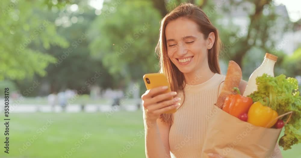 Cheerful young woman looking at smartphone screen and laughing. Beautiful millennial girl smiling while standing with her shopping in paper bag and using her phone. Concept of lifestyle Cheerful young woman looking at smartphone screen and laughing. Beautiful millennial girl smiling while standing with her shopping in paper bag and using her phone. Concept of lifestyle
