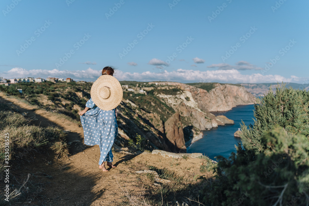 A girl in a blue dress in white polka dots and a hat stands on top of the sea, looking at the shoreline below