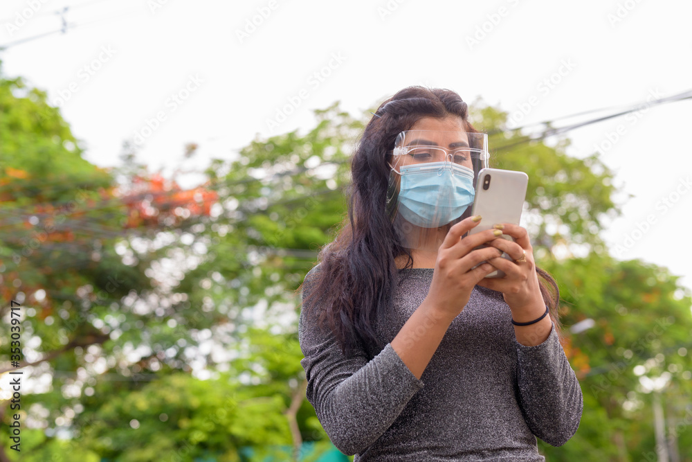Young Indian woman with mask and face shield using phone at the park outdoors Young Indian woman with mask and face shield using phone at the park outdoors