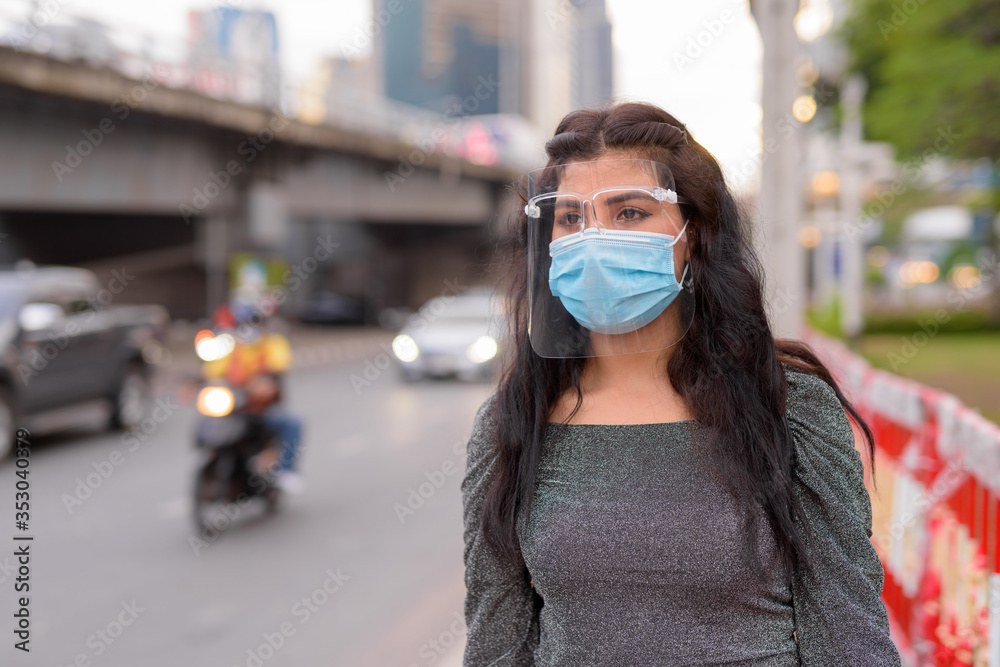 Fototapeta premium Young Indian woman with mask and face shield thinking in the city streets