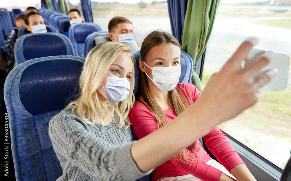 Fototapeta premium transport, tourism, road trip and people concept - young women or friends wearing face protective medical masks for protection from virus disease in travel bus taking selfie by smartphone