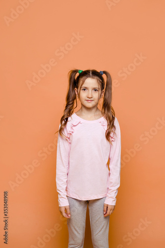 A girl with two ponytails stands on a pink background in daily clothes