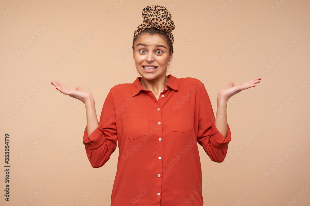 Confused young pretty brunette woman with headband grimacing her face while shrugging with raised palms, posing over beige background in red shirt and headband Confused young pretty brunette woman with headband grimacing her face while shrugging with raised palms, posing over beige background in red shirt and headband
