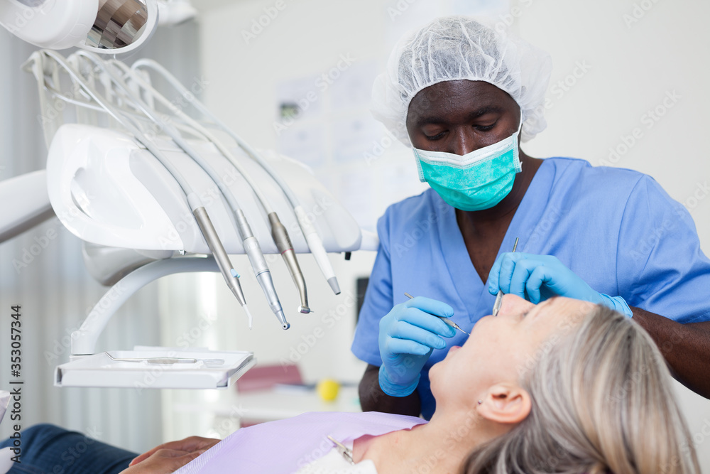 Dentist is treating female patient which is sitting in dental chair in clinic - obrazy, fototapety, plakaty 