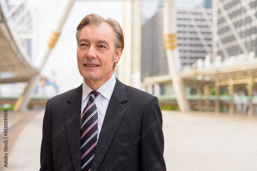 Happy mature handsome businessman in suit thinking at skywalk bridge in the city